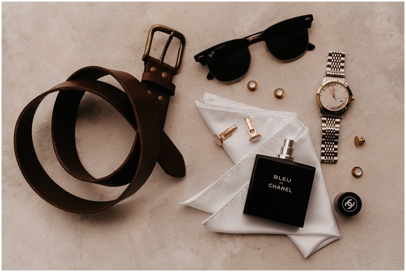 flat lay of groom wedding details including shoes, cufflinks, belt, watch, glasses, cologne, and pocket square